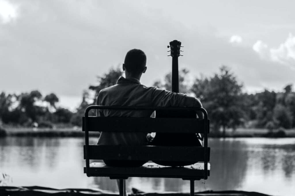 grayscale photo of man sitting beside guitar